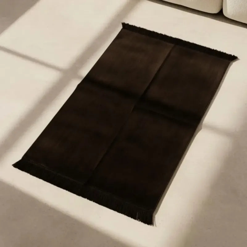 Een donkerbruine gebedskleed effen kleur met zachte textuur en franjes ligt plat op een lichte vloer