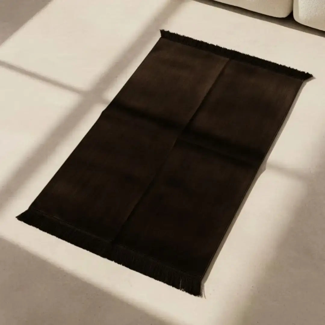 Een donkerbruine gebedskleed effen kleur met zachte textuur en franjes ligt plat op een lichte vloer