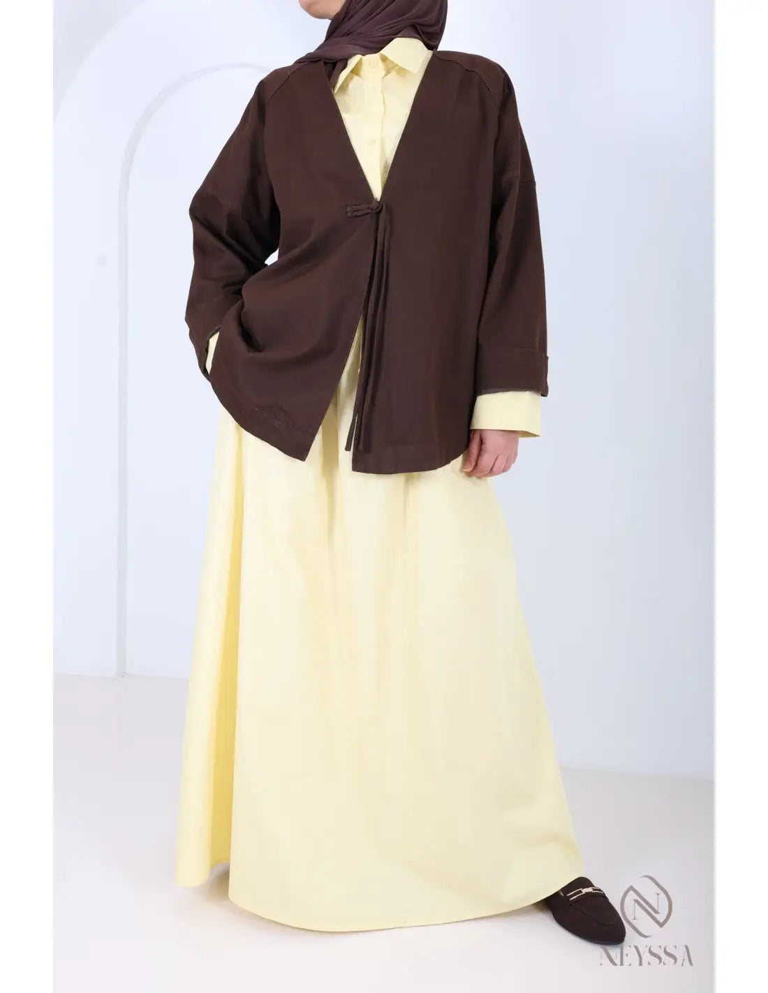 Een vrouw draagt een bruin kimono stijl jasje met voorkant sluiting en franjes over een geel jurkje