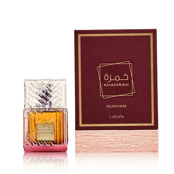 Een helder geslepen glazen parfumfles met verloop amber naar roze vloeistof en gouden folieetiket naast zijn bordeaux doosje van khamrah dukhan